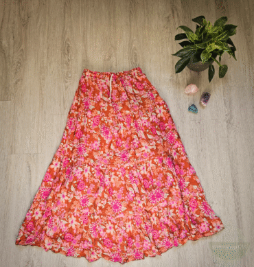 Langer Boho-Maxirock mit orangefarbenem Grundton und leuchtendem Blumenmuster in Pink und Weiß, ausgebreitet auf hellem Holzboden mit Zimmerpflanze und Heilsteinen daneben.