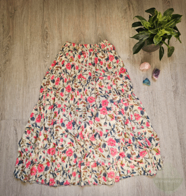 Boho Maxirock mit floralem Muster in Rosa, Blau und Braun auf cremefarbenem Hintergrund, ausgebreitet auf hellem Holzboden mit Zimmerpflanze und Edelsteinen daneben.