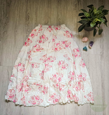 Langer cremeweißer Maxirock mit rosa Blumenprint und goldenen Pünktchen, ausgebreitet auf hellem Boden neben Heilsteinen und Pflanze