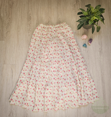 Langer Boho-Rock mit rosa Blumenmuster auf cremefarbenem Stoff, fließend ausgebreitet auf hellem Holzboden, daneben grüne Zimmerpflanze und Heilsteine.