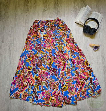 Langer Boho-Stufenrock in Königsblau mit buntem floralen Muster, ausgebreitet auf hellem Holz, daneben Buch, Amethyst und Kopfhörer – verträumtes Flatlay.