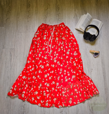 Langer roter Maxirock mit weißem Blumenmuster, auf einem Holzfußboden arrangiert, daneben ein offenes Buch, Kopfhörer, Amethyst und Räucherholz – Boho-Stil.