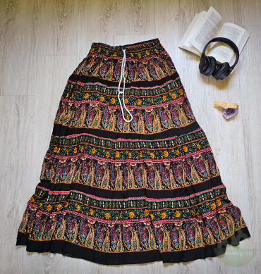 Handbedruckter schwarzer Maxirock mit buntem Paisley-Muster und Kordelzug, auf einem Holzboden liegend mit Kopfhörern und Buch.